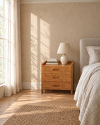 Honey bamboo bedside table with three drawers beside a linen bed in a British bedroom with a sash window