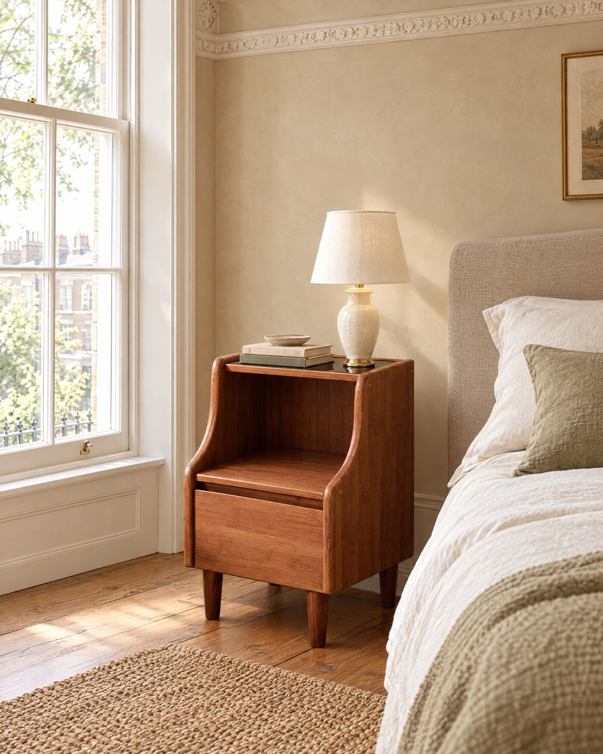 Walnut-tone bamboo bedside cabinet with curved sides, black top, open shelf and drawer beside a linen bed in a British bedroom with a sash window