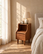 Walnut-tone bamboo bedside cabinet with curved sides, black top, open shelf and drawer with a ceramic lamp in morning light