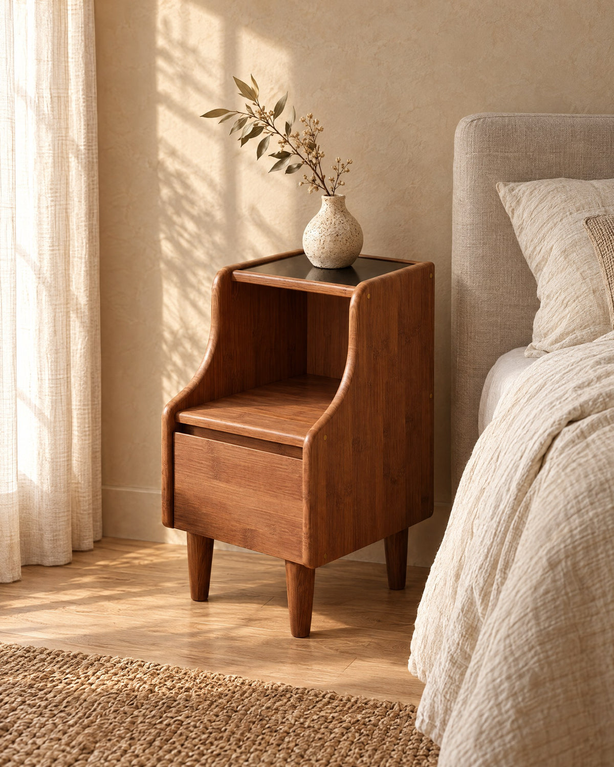 Three-quarter view of the walnut-tone bamboo bedside cabinet with curved sides, black top, open shelf and drawer