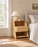 Bamboo bedside table with rattan side panels beside a linen bed in a British bedroom with a sash window
