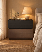 Modern bedside table of two stacked blocks, black wood grain over walnut tone, lamp switched on and an open book in the evening