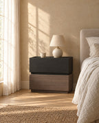 Modern bedside table of two stacked blocks, black wood grain over walnut tone with a ceramic lamp in morning light