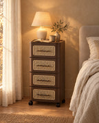 Brown rolling bedside chest with four cane-effect drawers and a lamp switched on in the evening