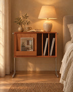 Cherry wood record storage bedside cabinet with chrome legs, lamp switched on and an open book in the evening