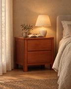Cherry-tone wooden bedside table with two handle-free drawers, lamp switched on and an open book in the evening