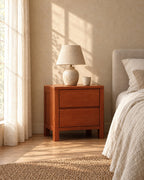 Cherry-tone wooden bedside table with two handle-free drawers with a ceramic lamp in morning light