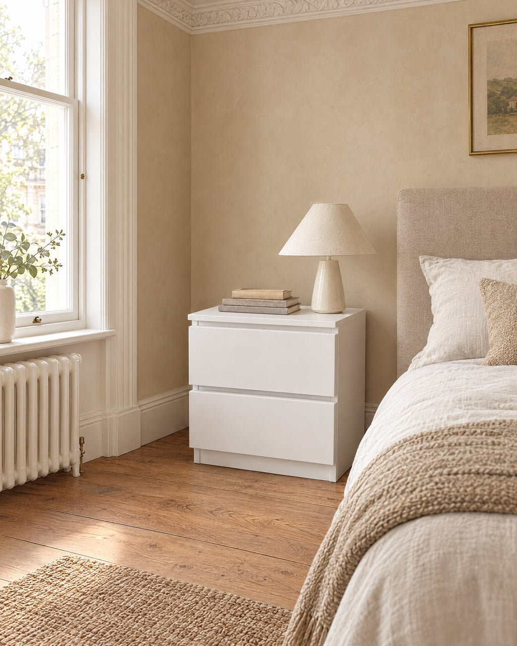 Compact white bedside table beside a linen bed in a bright British bedroom with a sash window
