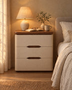Cream bedside table with three drawers and a walnut-tone top, a lamp switched on and an open book in the evening