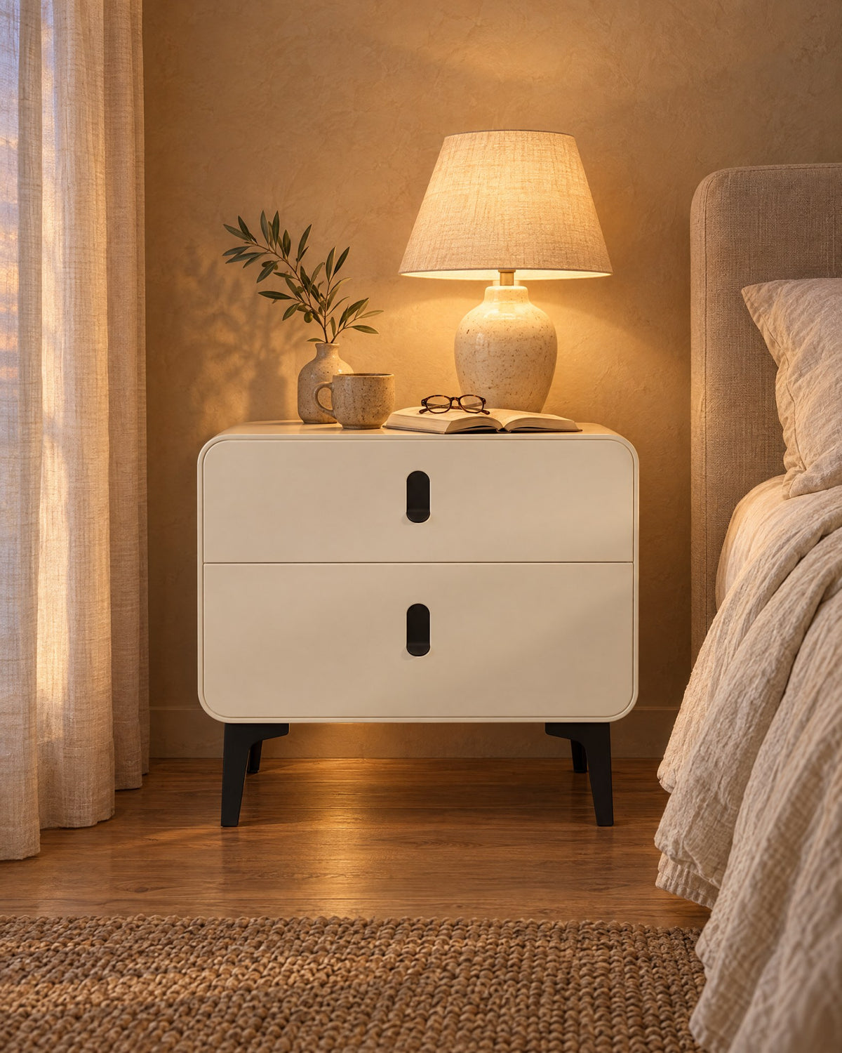 Cream bedside table with black metal legs, lamp switched on and an open book in the evening