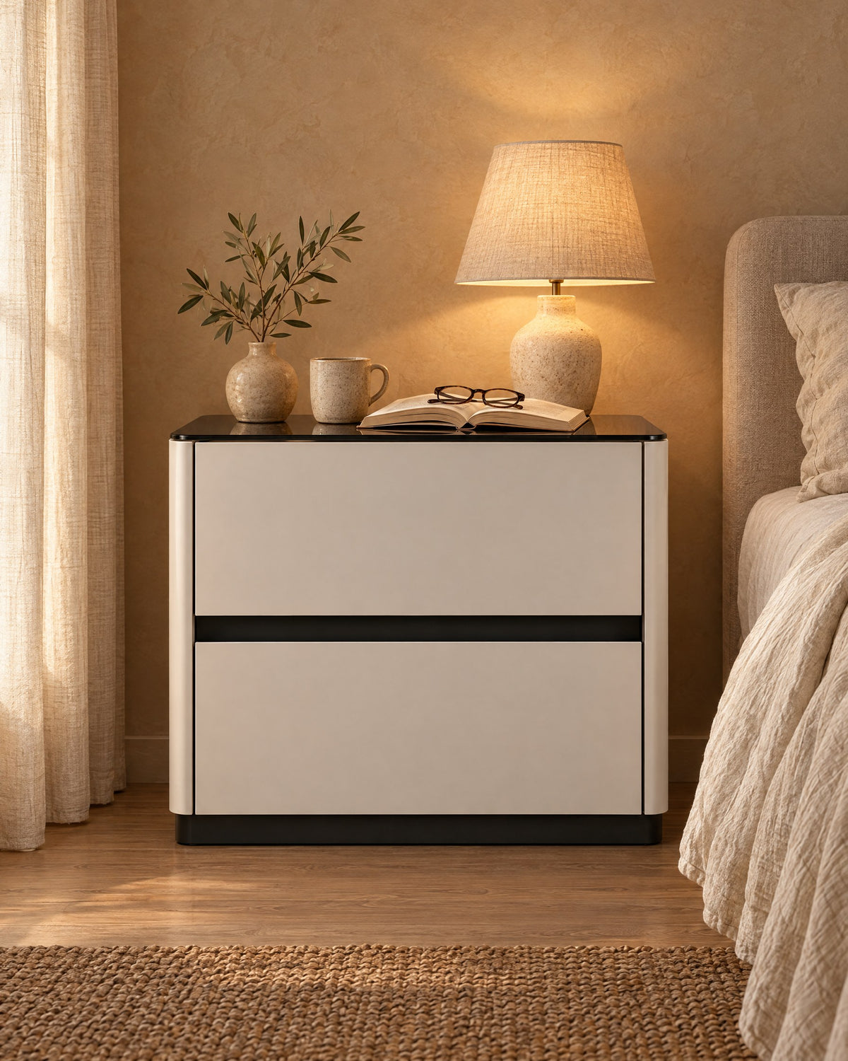 Cream bedside table with a glossy black top and two drawers, a lamp switched on and an open book in the evening