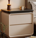 Front view of the cream bedside table with two drawers and a black top beside a bed