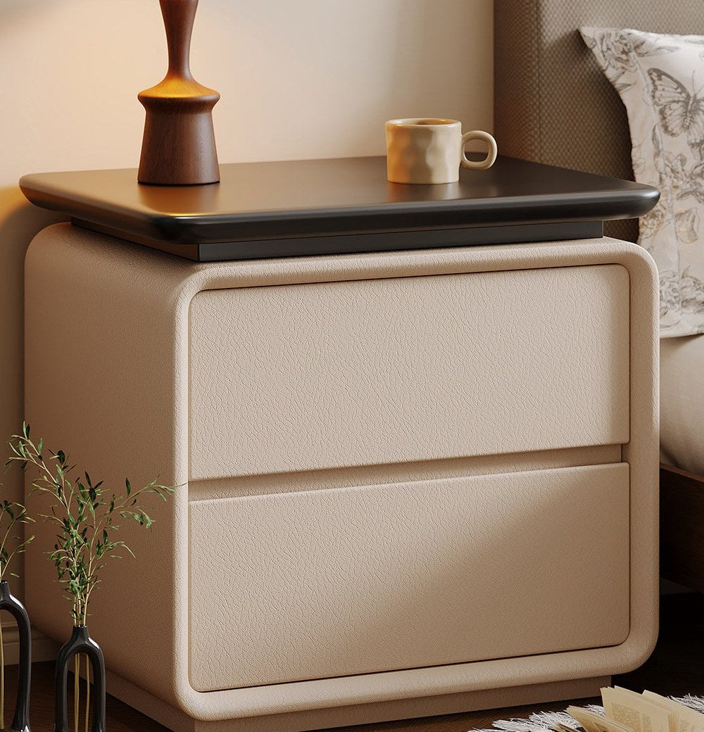 Front view of the cream bedside table with two drawers and a black top beside a bed