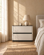 Cream bedside table with a glossy black top and two drawers with a ceramic lamp in morning light
