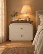 Cream bedside table with walnut-tone top and wooden knobs with a lamp switched on, an open book and reading glasses in the evening