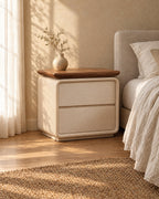 Three-quarter view of the cream bedside table with a walnut-tone top and two drawers beside a bed