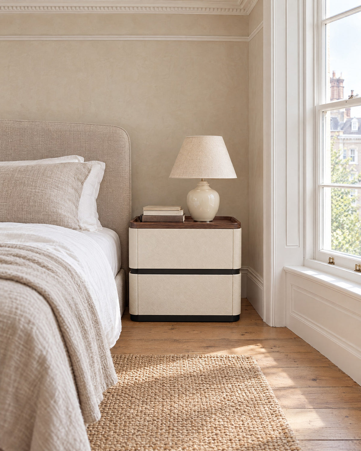 Cream bedside table with walnut tray top beside a linen bed in a British bedroom with a sash window