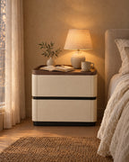 Cream bedside table with walnut tray top and two drawers, lamp switched on and an open book in the evening