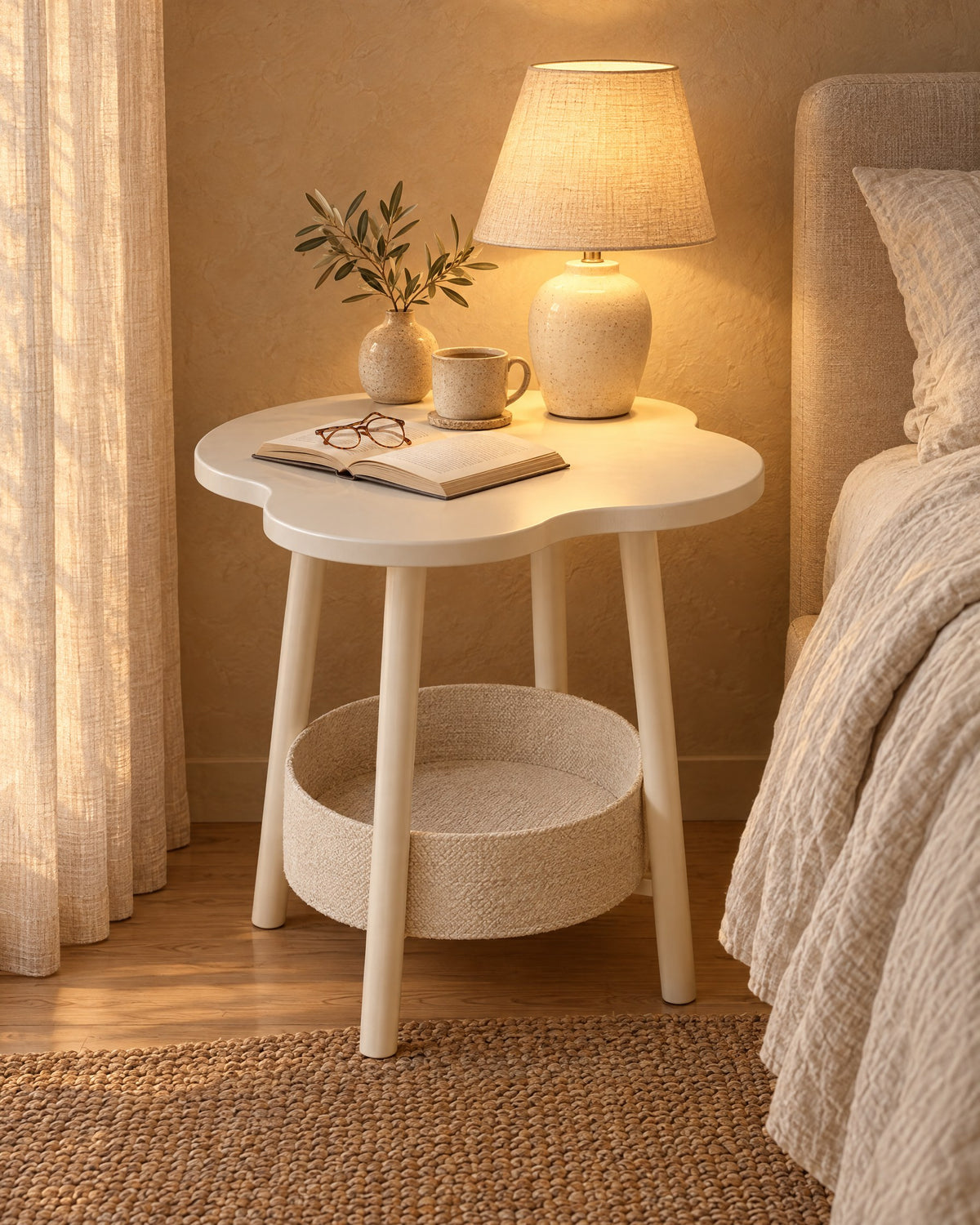 Cream flower-shaped bedside table with a storage basket with a lamp switched on, a book and reading glasses in the evening