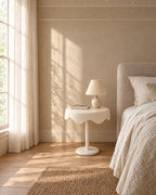 Cream round pedestal bedside table beside a linen bed in a bright British bedroom with a sash window