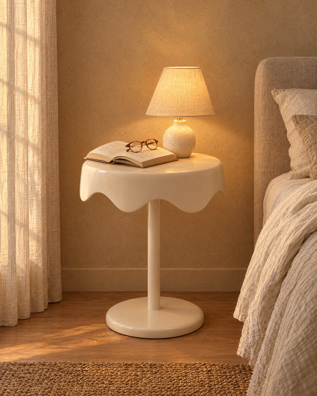 Cream round pedestal bedside table with a lamp switched on, an open book and reading glasses in the evening