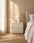 Cream rounded bedside table with two handle-free drawers with a ceramic lamp in morning light