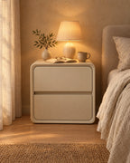 Cream rounded cube bedside table with a lamp switched on, an open book and reading glasses in the evening
