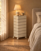 Cream seven-drawer swivel tower beside a bed with a lamp switched on in the evening