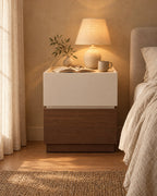 Cream and walnut two-tier bedside table with a lamp switched on and an open book in the evening