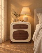 Cream bedside table with walnut drawer fronts and rounded feet, lamp switched on in the evening