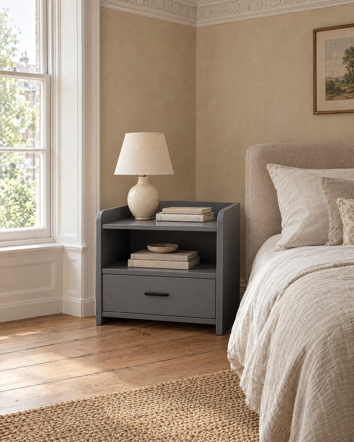 Grey bedside table with an open shelf and one drawer beside a linen bed in a bright British bedroom with a sash window