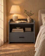 Grey bedside table with an open shelf and one drawer with a lamp switched on and an open book in the evening