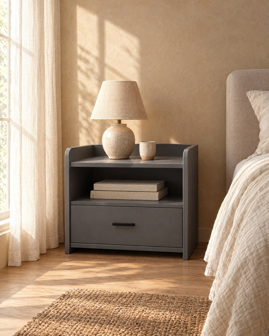 Grey bedside table with an open shelf and one drawer with a ceramic lamp in morning light