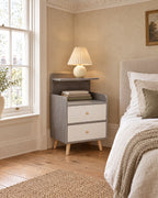 Grey bedside table with two white drawers and a raised shelf beside a linen bed in a bright British bedroom with a sash window