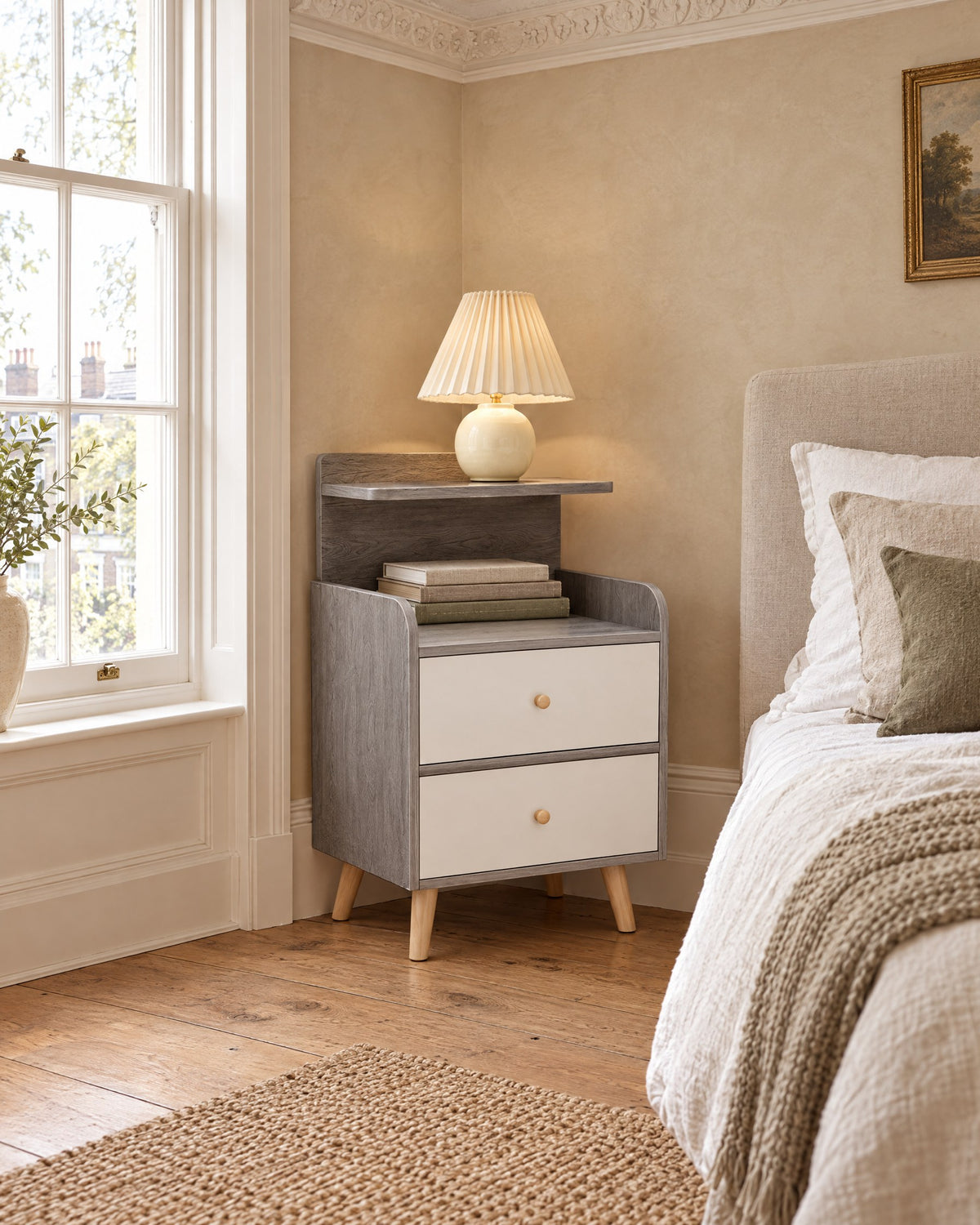 Grey bedside table with two white drawers and a raised shelf beside a linen bed in a bright British bedroom with a sash window