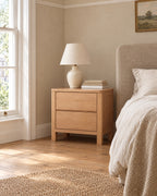 Honey wood bedside table with two drawers and square legs beside a linen bed in a British bedroom with a sash window
