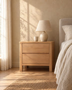 Honey wood bedside table with two drawers and square legs with a ceramic lamp in morning light