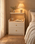 Light wood bedside table with two drawers and a floating shelf, lamp switched on and an open book in the evening