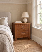 Lockable brown wooden bedside table with two drawers and scalloped rails beside a linen bed in a British bedroom with a sash window
