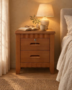 Lockable brown wooden bedside table with two drawers and scalloped rails, lamp switched on and an open book in the evening