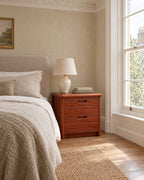Mahogany-tone wood bedside table with 2 drawers beside a linen bed in a bright British bedroom with a sash window
