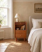 Narrow cherry wood bedside table beside a linen bed in a bright British bedroom with a sash window