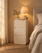 Narrow cream bedside table with three handle-free drawers with a lamp switched on, a book and reading glasses in the evening