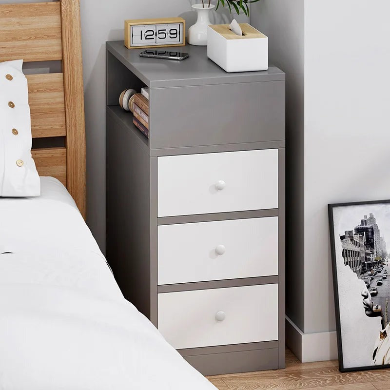 Narrow grey and white bedside table with three drawers beside a bed