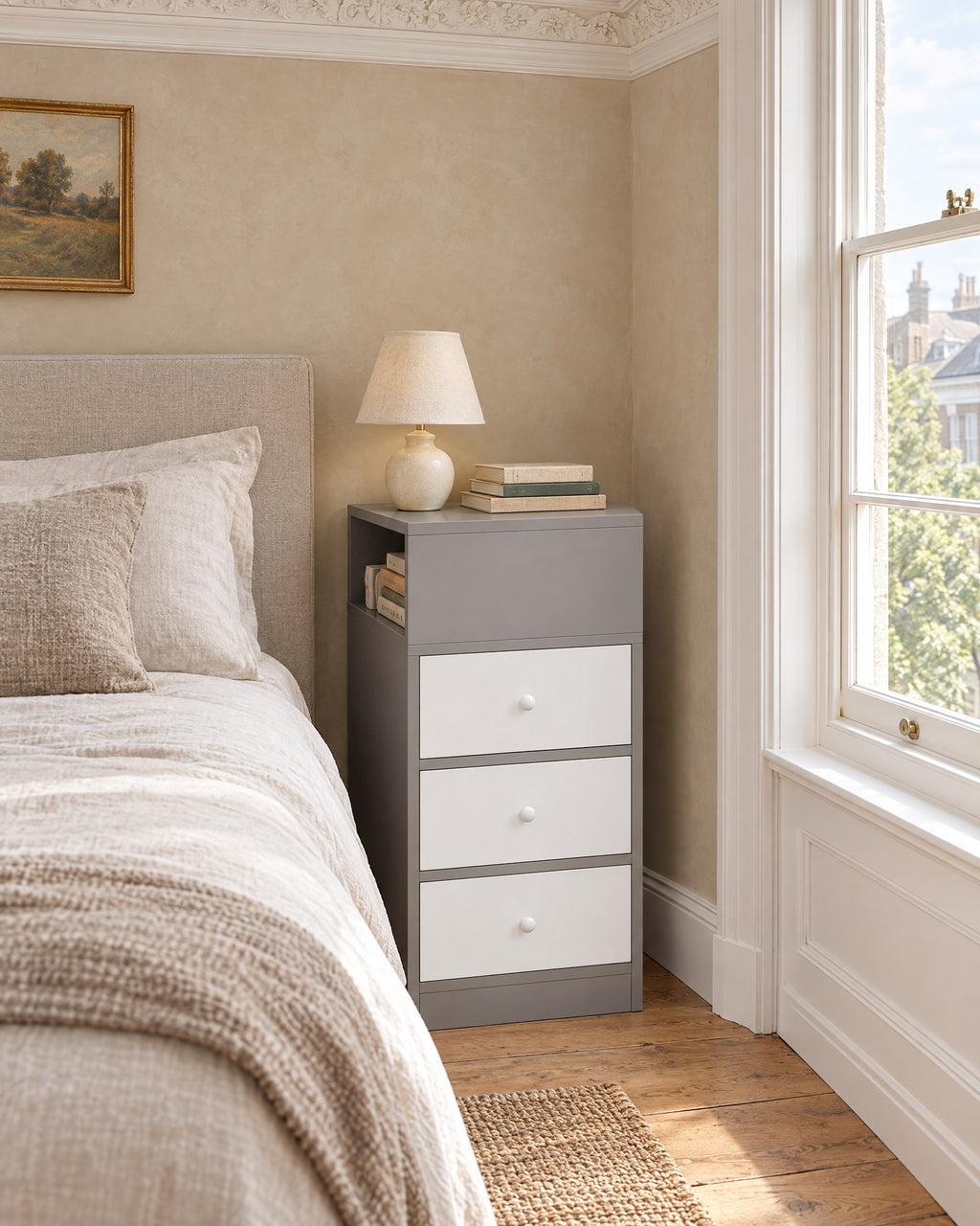Narrow grey and white bedside table beside a bed in a British bedroom with a sash window