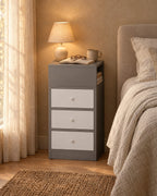 Narrow grey bedside table with three white drawers and a lamp switched on in the evening