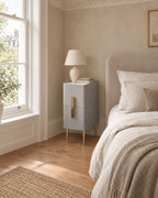 Narrow grey bedside table with a marble-effect top and gold legs beside a linen bed in a British bedroom with a sash window