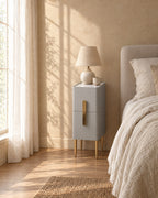 Narrow grey bedside table with a marble-effect top and gold legs with a ceramic lamp in warm morning light