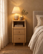 Narrow natural wood bedside table with a lamp switched on, a book and reading glasses in the evening