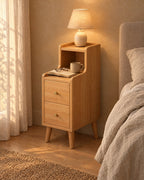 Narrow oak bedside table with a lamp switched on, an open book and glasses in the evening